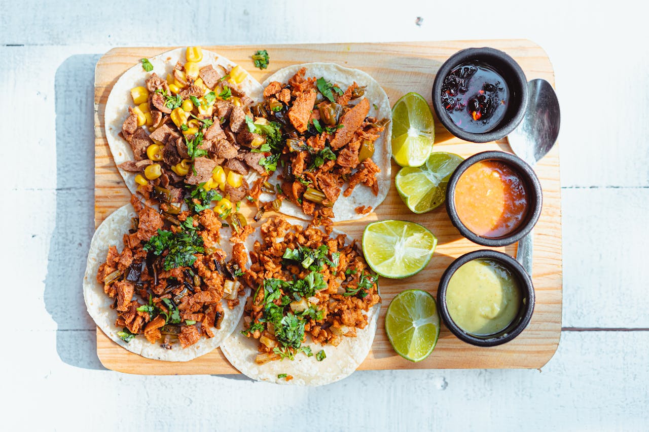 Colorful vegan tacos with lime slices and dipping sauces on a wooden tray, shot in bright light.